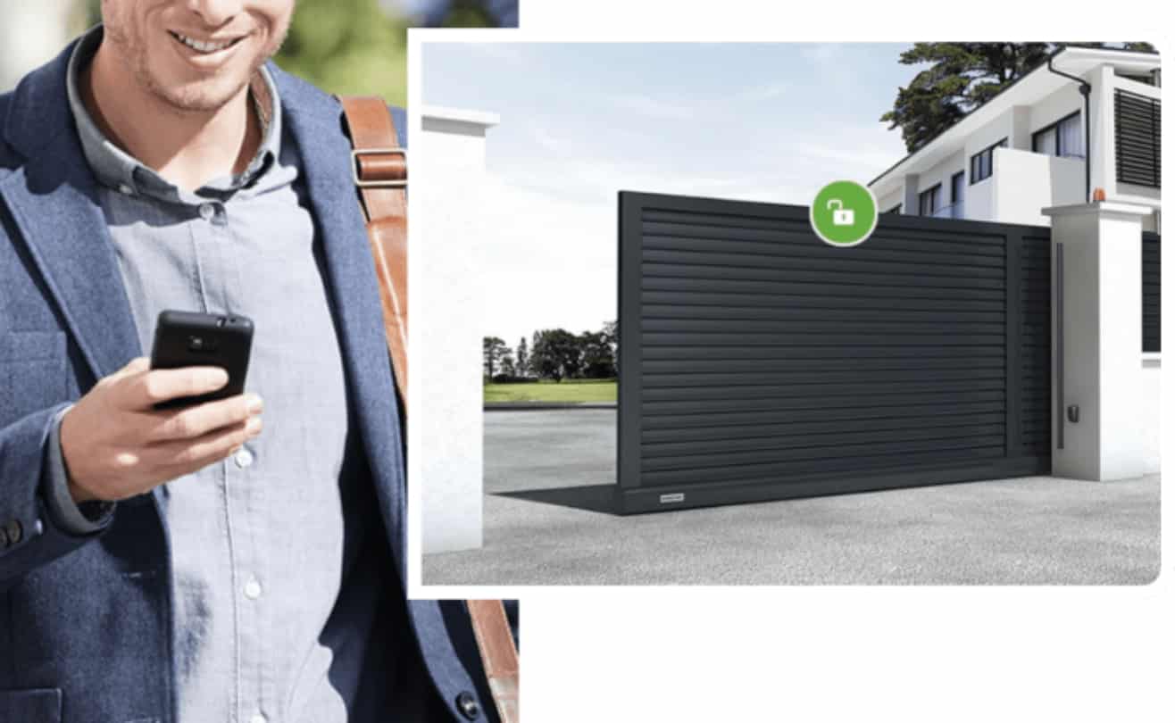 En mand i en blå blazer holder en smartphone og smiler. Ved siden af ham er en stor moderne sort port foran et hus, med et ulåst hængelåsikon over sig, der viser den praktiske indgang, der sikres og styres via det automatiserede smartphonesystem.