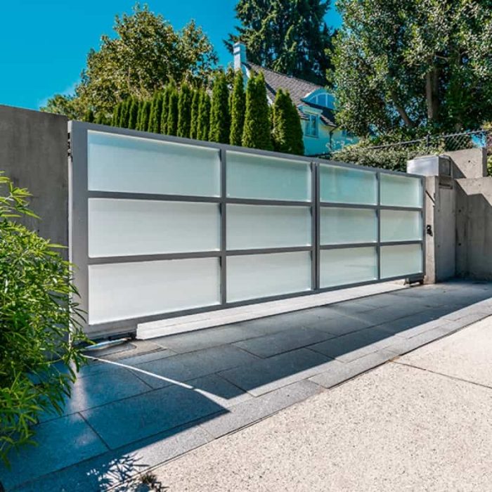 En moderne indkørselsport lavet af matte glaspaneler og metalrammer er installeret ved indgangen til et hjem. Den motoriserede port, udstyret med en ismartgate LITE - Fjernstyret Låge & portåbner via. Din Smartphone eller PC - (Max 1 tilslutning af låge eller port), kan styres via smartphone eller PC. Beliggende på en betonindkørsel, er det omgivet af frodige grønne områder og store træer, med huset synligt i baggrunden.