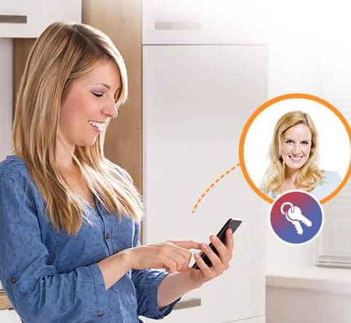 En smilende kvinde i en blå skjorte bruger sin smartphone i et lyst køkken, og hendes tankeboble viser en anden smilende kvinde med et nøgleikon. Hun diskuterer sandsynligvis bekvemmeligheden ved en ismartgate LITE - Fjernstyret Låge & portåbner via. Din smartphone eller pc til fjernadgang til port eller garageport.