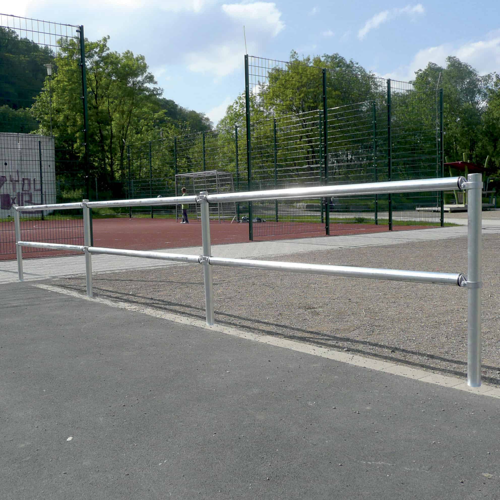En sportsbane med hegn og målstolpe i baggrunden. Banen er omgivet af et metalrækværk med et brolagt område i forgrunden og træer i det fjerne. Himlen er delvist overskyet, typisk for mange sportspladser.