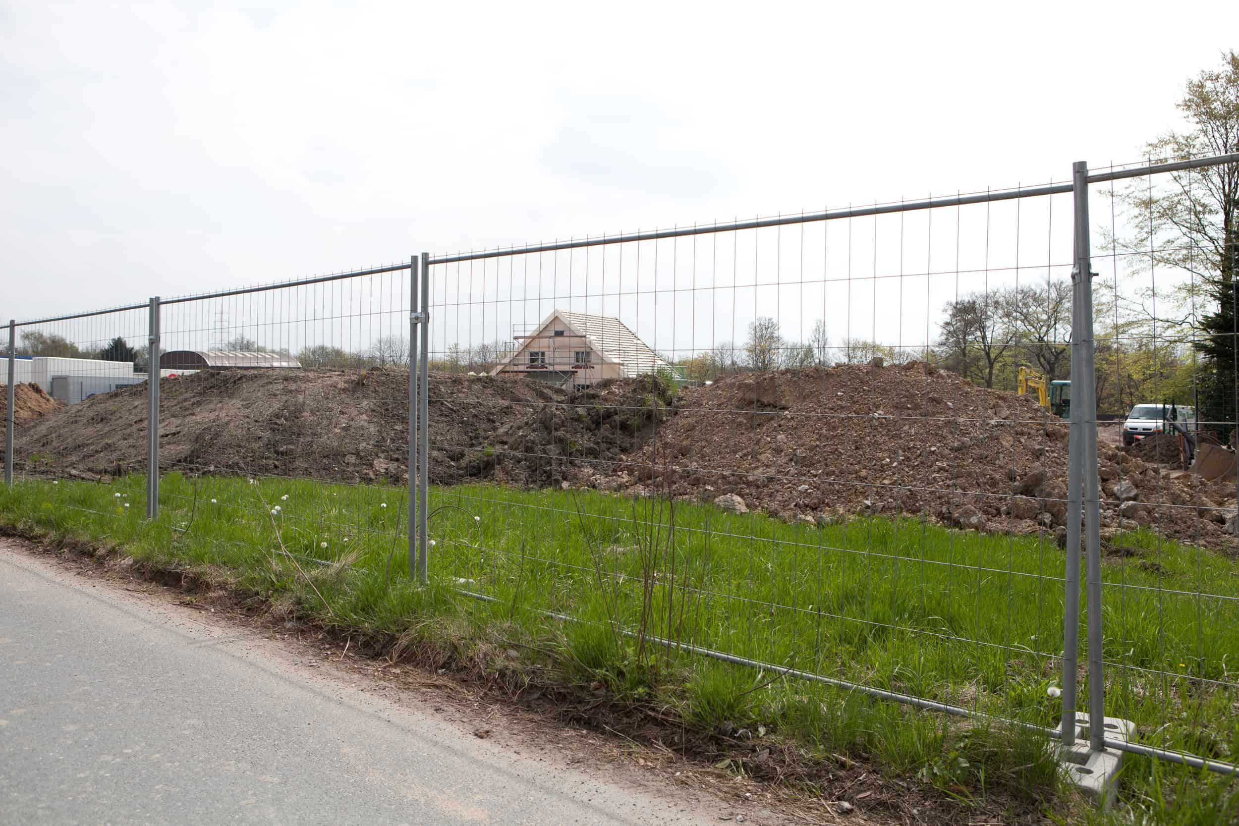 En byggeplads er omsluttet af en mobil hegn. Området indeholder bunker af snavs og byggematerialer. I baggrunden ses en delvist opført bygning med skrå tag. Forgrunden viser en vej og et græsareal. Træer og andre strukturer omgiver stedet.