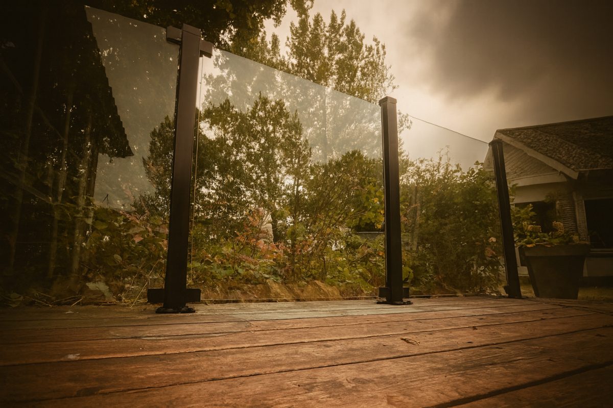 En lav vinkel på en træterrasse med glasrækværk, der reflekterer træer og planter udenfor. Himlen ovenover er overskyet, og en del af et hus med skråt tag ses til højre.