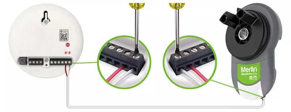 Et billede, der viser en ledningsproces, der forbinder en røgdetektor til en Merlin standby batteri backup enhed. Forbindelsen involverer fastgørelse af ledninger i en klemrække ved hjælp af skruer. Praktisk indgang funktioner fremhæver komponenterne med indsatte visninger, der detaljerer ledningstrinnene.