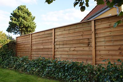 Et plankeværk af træ med vandrette planker står i en baghave med grøn vedbend, der vokser i bunden. I baggrunden er et træ og taget af et hus delvist synligt under en blå himmel med nogle skyer.