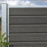 Et nærbillede af en "Komposit Hegn med galvaniseret stålstolper & u-skinner - Type 1" med grå vandrette lameller og metalstolper. En lille grøn plante ved siden af hegnet fremhæver dets slanke design, mens delvist overskyet himmel kaster bløde skygger på denne stilfulde udendørsfunktion.