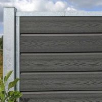 Et nærbillede af en "Komposit Hegn med galvaniseret stålstolper & u-skinner - Type 1" med grå vandrette lameller og metalstolper. En lille grøn plante ved siden af hegnet fremhæver dets slanke design, mens delvist overskyet himmel kaster bløde skygger på denne stilfulde udendørsfunktion.