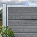 I en have står en grå trætekstureret "Komposit Hegn med galvaniseret stålstolper & u-skinner - Type 1" med en bladgrøn plante i bunden og en overskyet himmel i baggrunden.