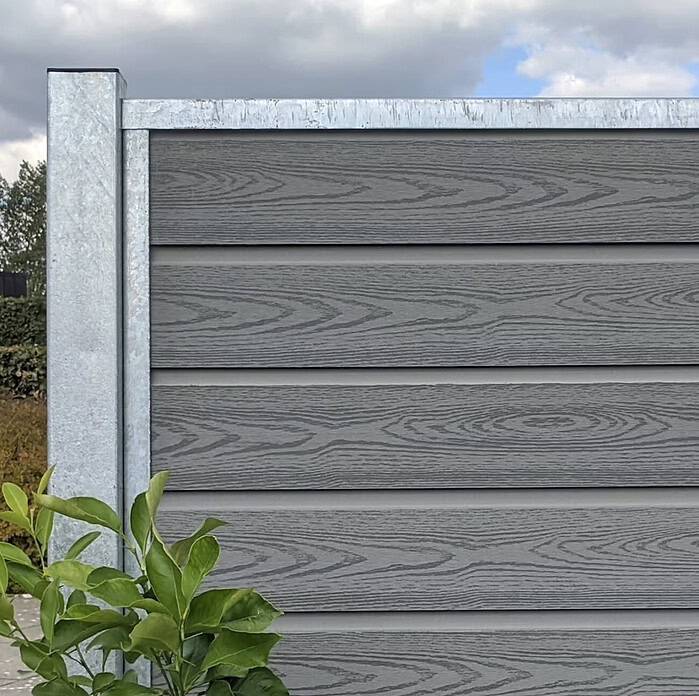 I en have står en grå trætekstureret "Komposit Hegn med galvaniseret stålstolper & u-skinner - Type 1" med en bladgrøn plante i bunden og en overskyet himmel i baggrunden.