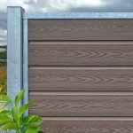 Mod den overskyede himmel ligner et hegn med vandrette lameller og metalstolper Komposit Hegn med galvaniseret stålstolper & u-skinner - Type 1. I forgrunden giver grønne blade kontrast, der problemfrit blander sig for en diskret, men slående scene.