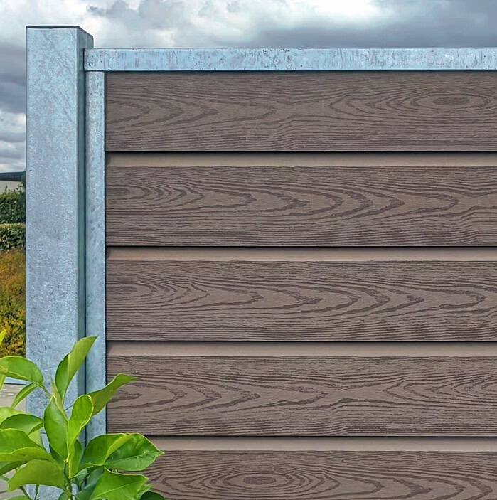 Mod den overskyede himmel ligner et hegn med vandrette lameller og metalstolper Komposit Hegn med galvaniseret stålstolper & u-skinner - Type 1. I forgrunden giver grønne blade kontrast, der problemfrit blander sig for en diskret, men slående scene.