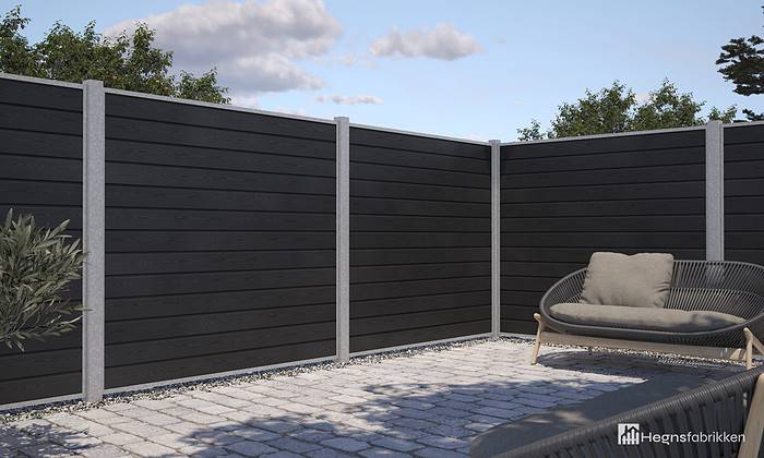 Et moderne udendørs terrasseområde med slankt sort vandret lamelhegn lavet af Komposit Hegn med galvaniseret stålstolper & u-skinner - Type 1 og grå støttestolper. Terrassen har en potteplante til venstre og en polstret sofa med en taupe-pude til højre, alt sammen sat på lysegrå stenbelagt jord. Himlen er delvist overskyet.