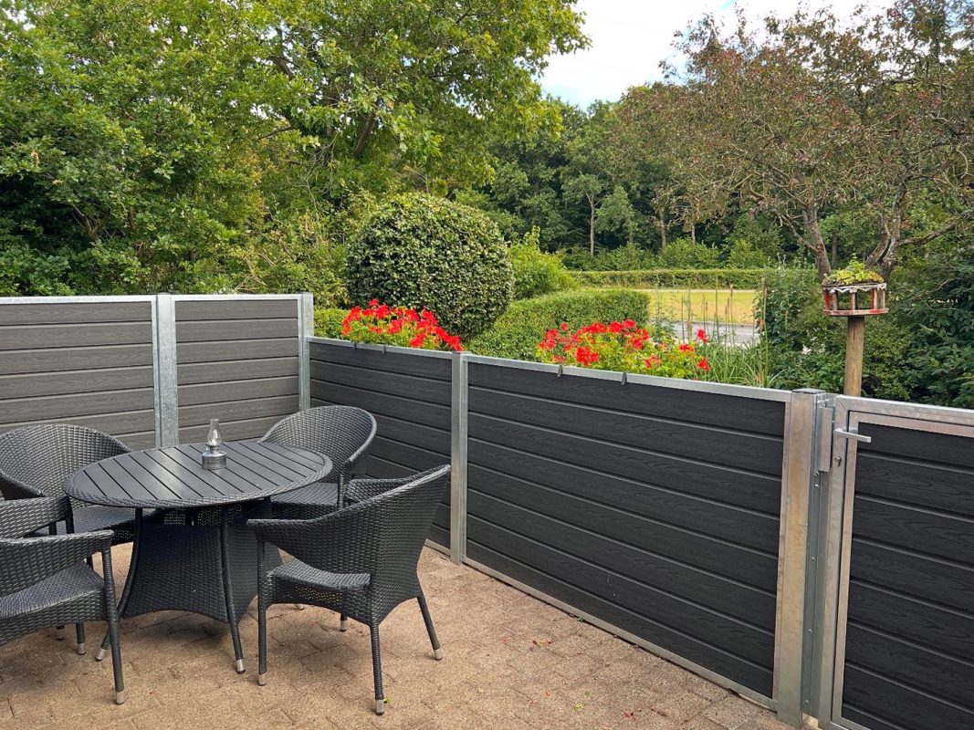 En terrasse med et rundt sort bord og fire flettede stole er omgivet af et Komposit Hegn med varmgalvaniseret stålstolper & u-skinner - Type 1. Røde blomster står langs hegnet med træer og grønne områder i baggrunden.