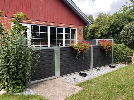 Et murstenshus med store hvide vinduer og en Komposit Hegn med varmgalvaniseret stålstolper & u-skinner - Type 1. Røde blomster i krukker giver farve, og grønne buske og en græsplæne afgrænser grusstien ved siden af hegnet.