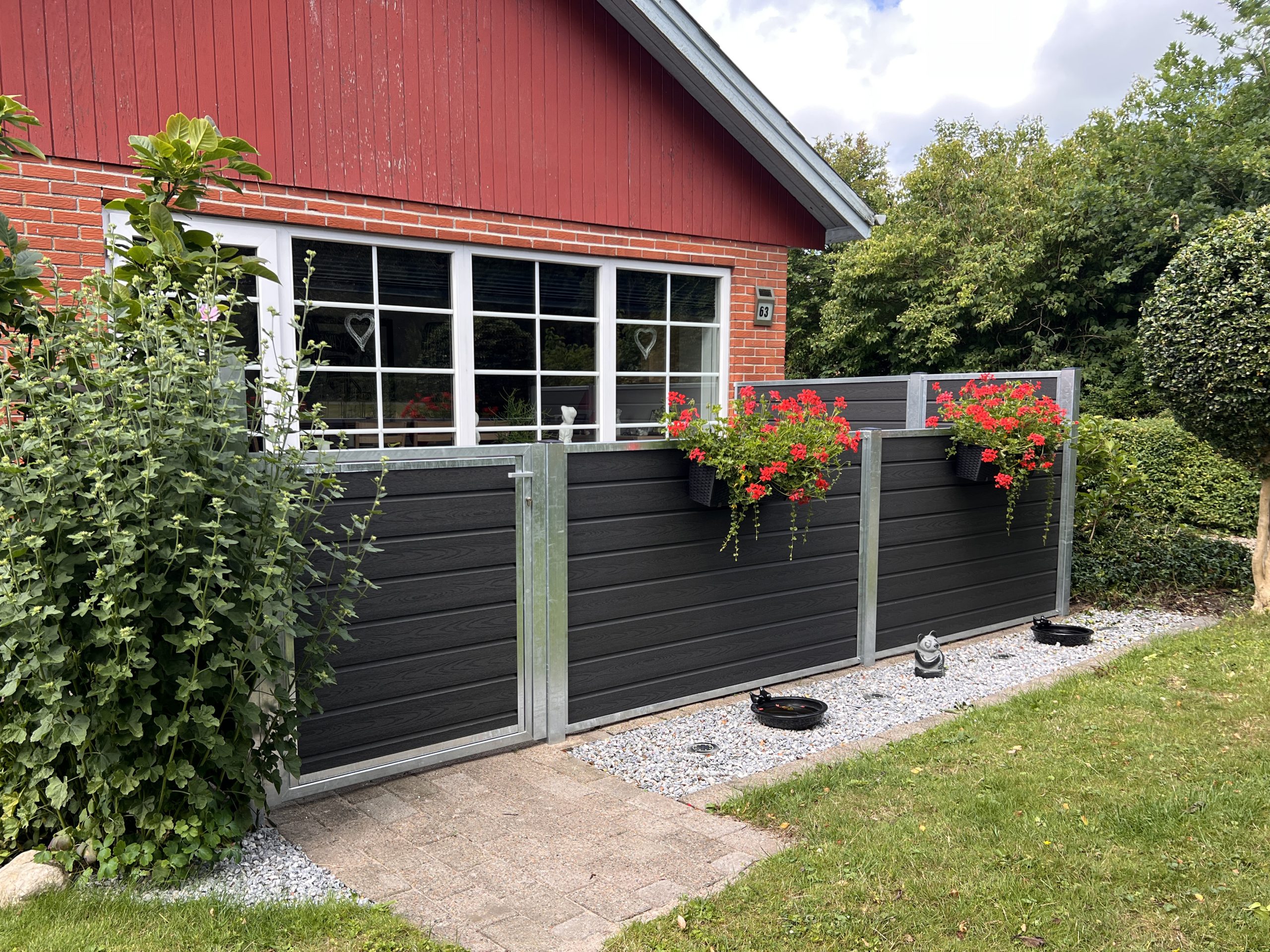 Et murstenshus med store hvide vinduer og en Komposit Hegn med varmgalvaniseret stålstolper & u-skinner - Type 1. Røde blomster i krukker giver farve, og grønne buske og en græsplæne afgrænser grusstien ved siden af hegnet.