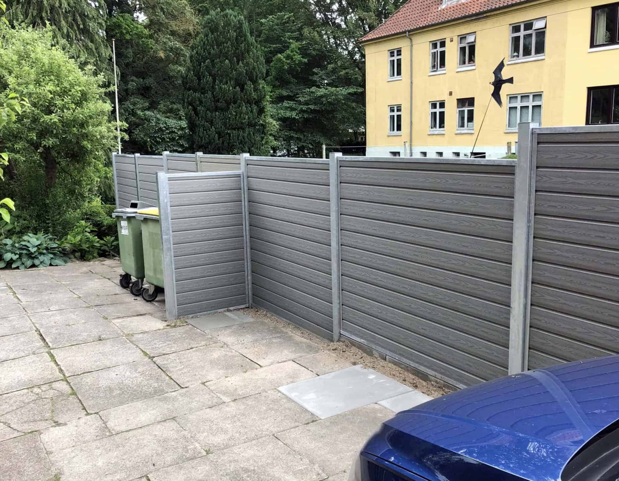 Komposit Hegn med varmgalvaniseret stålstolper & u-skinner - Type 1 adskiller et asfalteret område med skraldespande og en blå bil fra en have, med en gul bygning og træer synlige i baggrunden.