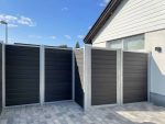 Et moderne sort/hvidt hegn med vandrette paneler af Komposit Hegn med galvaniseret stålstolper & u-skinner - Type 1 ses i hjørnet af et brolagt område uden for et hvidt hus. Himlen er klar med enkelte skyer.