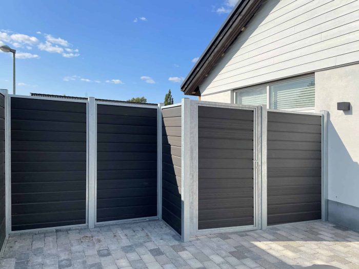 Et moderne sort/hvidt hegn med vandrette paneler af Komposit Hegn med galvaniseret stålstolper & u-skinner - Type 1 ses i hjørnet af et brolagt område uden for et hvidt hus. Himlen er klar med enkelte skyer.