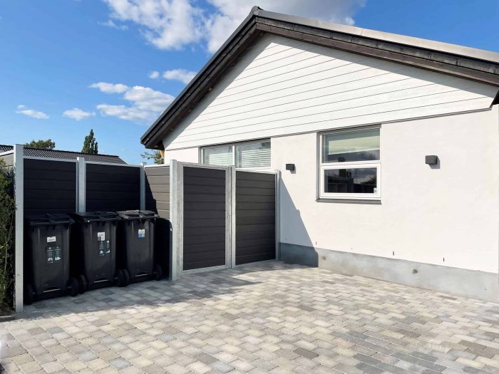 Et moderne hus med hvid facade og gråt tag under en blå himmel. Indkørslen er lavet af lysegrå sammenlåsende brosten. Til venstre står tre sorte skraldespande opstillet ved siden af en lækker sort og grå Komposit Hegn med galvaniseret stålstolper & u-skinner - Type 1. Huset har to vinduer og to væghængte lamper.