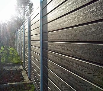 Nærbillede af et langt, mørkt træhegn med Komposit Hegn med galvaniseret stålstolper & u-skinner - Type 1, set fra en vinkel under stærkt sollys. Hegnet strækker sig i det fjerne med træer synlige i baggrunden i venstre side. Nogle græs- og havearealer er foran.