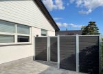 Et moderne hus med lys beklædning og en række af tre-rude vinduer står elegant. Ved siden af er en Komposit Hegn med galvaniseret stålstolper & u-skinner - Type 1, med mørke vandrette paneler, der kaster skygger på en brolagt terrasse. I baggrunden kan man skimte et nabohus og en delvist overskyet himmel.