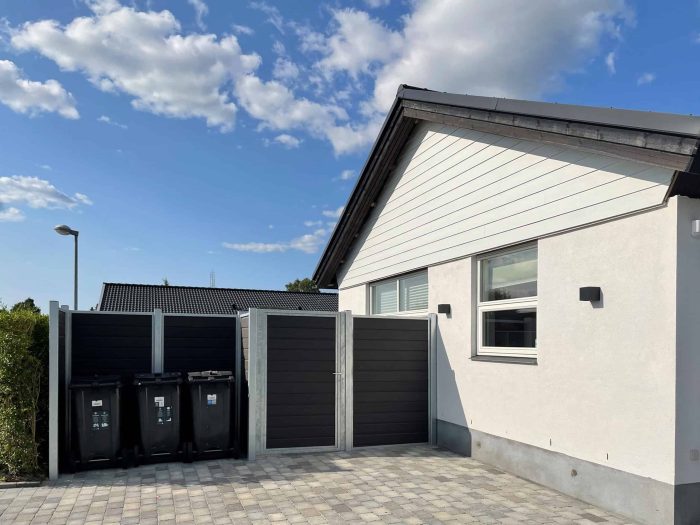 Et moderne et-plans hus med hvide beklædninger og et gråt tag under en klar blå himmel. Huset har en brolagt indkørsel og til venstre side et indhegnet område med tre sorte skraldespande, omkranset af en elegant Komposit Hegn med galvaniseret stålstolper & u-skinner - Type 1.