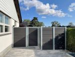 Et moderne hus udvendigt med gråt sidespor har et indhegnet område lavet med Komposit Hegn med galvaniseret stålstolper & u-skinner - Type 1, med mørke lodrette trælameller og metalrammer. Hegnet inkluderer en port og er placeret på stenbelægning, med buske til højre og en klar blå himmel oversået med skyer.