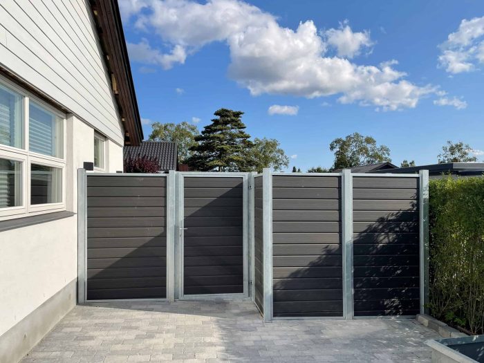 Et moderne hus udvendigt med gråt sidespor har et indhegnet område lavet med Komposit Hegn med galvaniseret stålstolper & u-skinner - Type 1, med mørke lodrette trælameller og metalrammer. Hegnet inkluderer en port og er placeret på stenbelægning, med buske til højre og en klar blå himmel oversået med skyer.