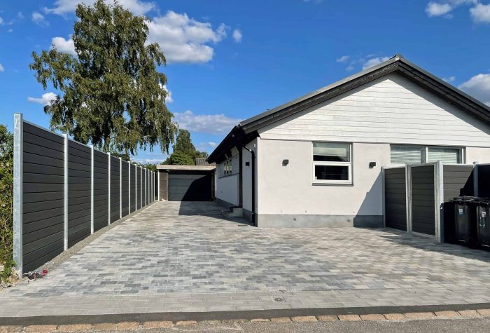 Et moderne en-etagers hus med hvide ydervægge og mørk tagshingles har en rummelig indkørsel lavet af sammenlåsende grå mursten, der fører til en fritliggende garage. Ejendommen er omkranset af et højt mørkt hegn opført af Komposit Hegn med galvaniseret stålstolper & u-skinner - Type 1. Et træ og en klar blå himmel er i baggrunden.