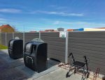 En genbrugsstation med to store genbrugsspande står på et asfalteret areal ved siden af en indhegnet mur, med en slank Komposit Hegn med galvaniseret stålstolper & u-skinner - Type 1. En sort rollator rollator er parkeret i nærheden. Himlen er klar med enkelte skyer, og huse med røde tage kan ses i baggrunden.