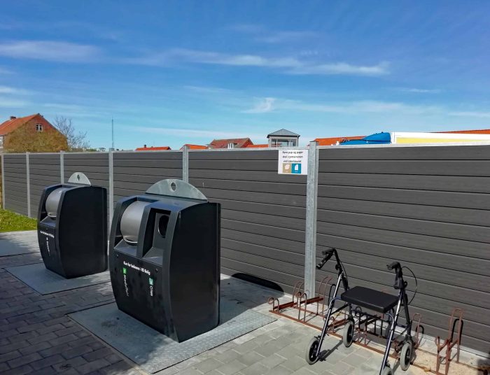 En genbrugsstation med to store genbrugsspande står på et asfalteret areal ved siden af en indhegnet mur, med en slank Komposit Hegn med galvaniseret stålstolper & u-skinner - Type 1. En sort rollator rollator er parkeret i nærheden. Himlen er klar med enkelte skyer, og huse med røde tage kan ses i baggrunden.