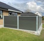 Et moderne hus med mørkt tegltag og ydre i beige mursten har et indhegnet område med grå paneler og metalstolper. Komposit Hegn med galvaniseret stålstolper & u-skinner - Type 1 står på en hævet betonplatform, og gården ud over hegnet har en velholdt grøn græsplæne under en overskyet himmel.