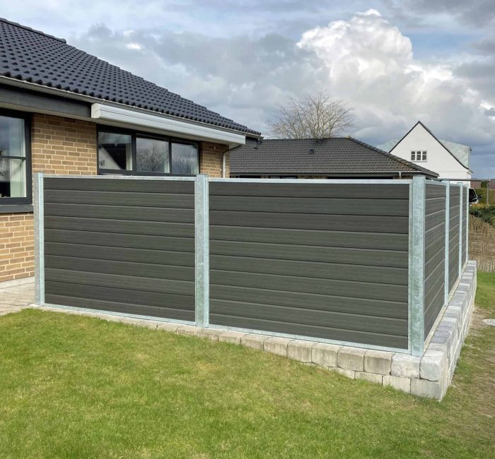 Et moderne hus med mørkt tegltag og ydre i beige mursten har et indhegnet område med grå paneler og metalstolper. Komposit Hegn med galvaniseret stålstolper & u-skinner - Type 1 står på en hævet betonplatform, og gården ud over hegnet har en velholdt grøn græsplæne under en overskyet himmel.