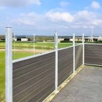 Et moderne udendørsområde med Komposit Hegn med klartglas - Inkl. galvaniseret stålstolper u-skinner og træpaneler hegn, med udsigt over et græsklædt landskab med bygninger i det fjerne under en delvist overskyet himmel. Området omfatter klinkegulv og hegn understøttet af galvaniseret stålstolper.