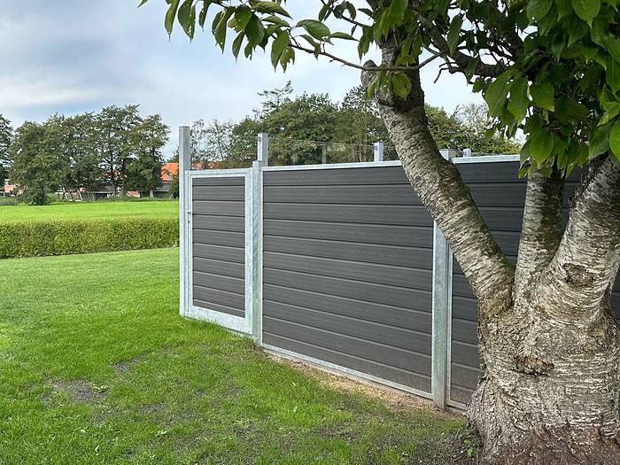 Et stort træ står i forgrunden på en græsplæne, ved siden af en høj, mørkfarvet Komposit Hegn med klartglas - Inkl. galvaniseret stålstolper u-skinner. Hegnet strækker sig på tværs af billedet og omslutter delvist et område. Frodigt grønt og en række træer er synlige i baggrunden.