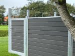 Et nærbillede af et moderne, gråt hegn sammensat af vandrette trælameller og galvaniseret stålstolper med metalramme, med låge. Dette er Komposit Hegn med klartglas - Inkl. galvaniseret stålstolper u-skinner model. Hegnet er placeret på en frodig grøn græsplæne med nogle træer i baggrunden og en træstamme delvist synlig i højre side, mens himlen er overskyet.