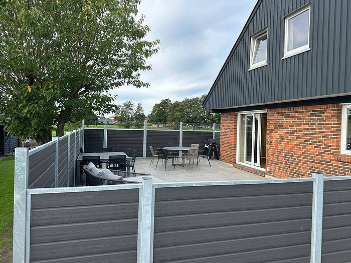 Et moderne hus med delvist muret, delvis mørkt beklædning udvendigt har et indhegnet terrasseområde med Komposit Hegn med klartglas - Inkl. galvaniseret stålstolper u-skinner. Terrassen har udendørsmøbler, herunder stole, borde og et hjørnesofasæt. Galvaniseret stålstolper tilføjer holdbarhed til indhegningen. Et træ står på den ene side og grønt er synligt i baggrunden.