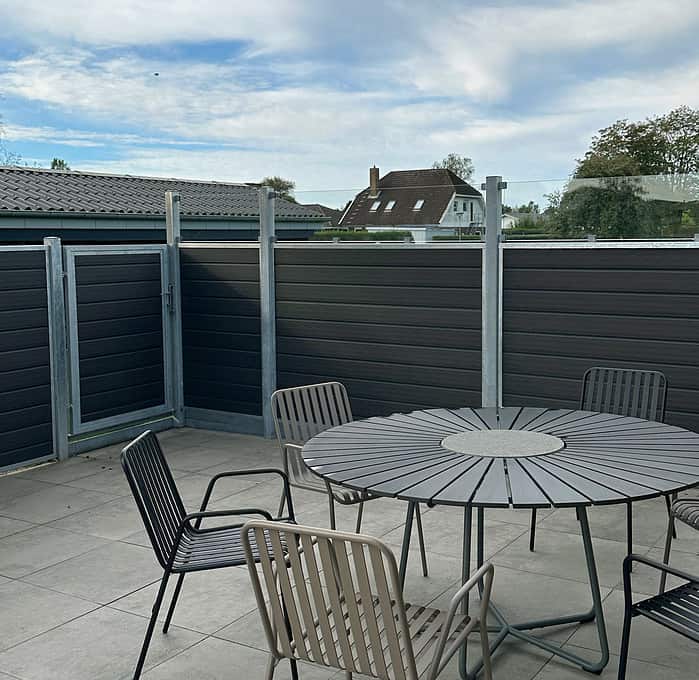 En tagterrasse har et metalbord og flere stole. Området er omsluttet af Komposit Hegn med klart glas paneler, herunder galvaniseret stålstolper u-skinner for solid støtte. Delvis skyet himmel pryder baggrunden, hvor bygninger og træer kan ses. Terrassegulvet er sammensat af lyse fliser.