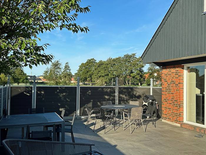 Et gårdhaveområde med flere metalstole og -borde, en grill og en glasdør, der fører til en murstens- og mørk træbygning. Pladsen er omsluttet af Komposit Hegn med klartglas - Inkl. galvaniseret stålstolper u-skinner til støtte, mens træer indrammer scenen mod en klar blå himmel.