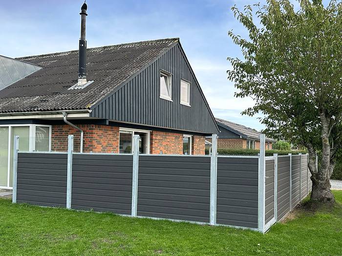 Et hjørne af et moderne hus med mørkegrå og murstens ydre, skrånende tag og høj skorsten. Huset har en wraparound Komposit Hegn med klartglas - Inkl. galvaniseret stålstolper u-skinner og en grøn græsplæne. Der er et træ nær hegnet og klar himmel i baggrunden.