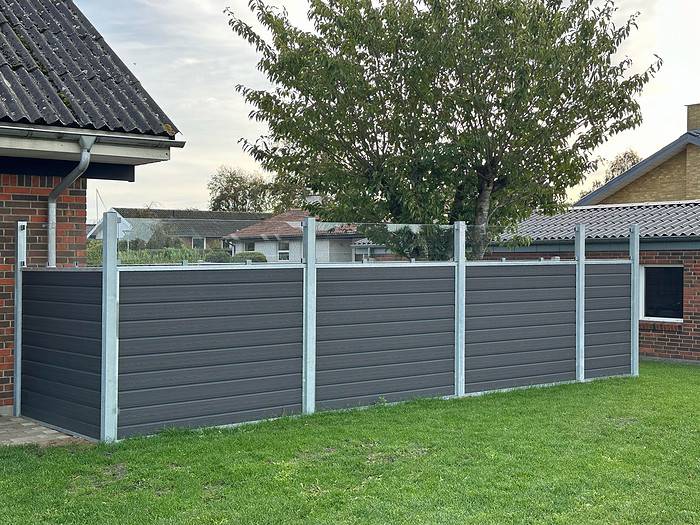 Et specialfremstillet privat hegn, Komposit Hegn med klartglas - Inkl. galvaniseret stålstolper u-skinner, med mørkegrå vandrette paneler omslutter et udendørsareal ved siden af et murstenshus. Dette stilfulde hegn er toppet med klare glaspaneler og sidder på en velplejet grøn græsplæne, komplet med et træ og andre huse synlige i baggrunden.