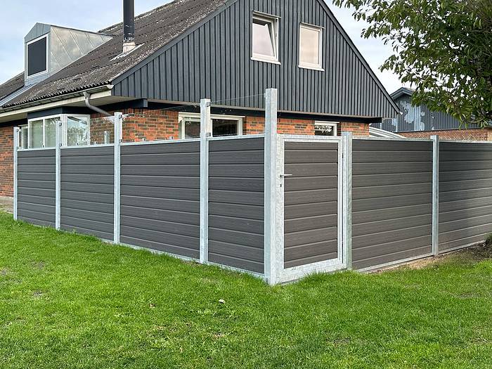 Et moderne hus med skrå tag med både mursten og mørk beklædning. Gården er omsluttet af et højt, gråt Komposit Hegn med klartglas - Inkl. galvaniseret stålstolper u-skinner og en låge. Græsset er grønt, og der er et træ til højre. Himlen er overskyet.