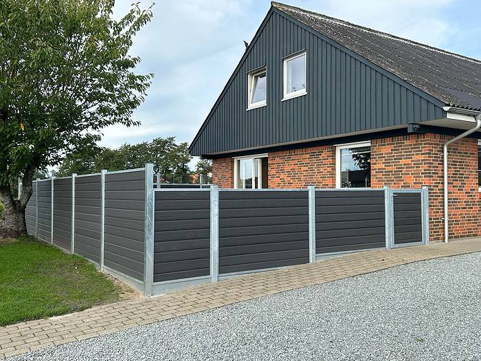 Et hus med muret underfacade og mørkegrå overfacade har en mørkegrå Komposit Hegn med klartglas - Inkl. galvaniseret stålstolper u-skinner. Det indhegnede område omfatter grusbelægning og grønt, med træer synlige i baggrunden under en delvist overskyet himmel.