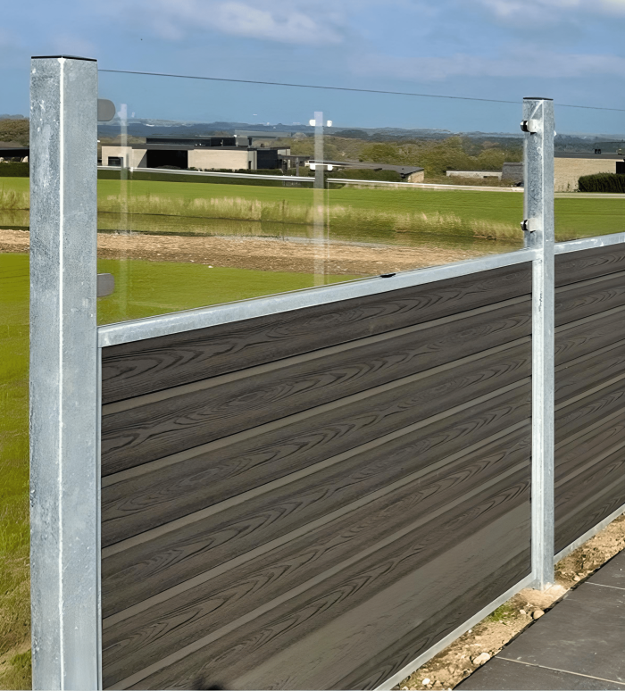 Et nærbillede af Komposit Hegn med klartglas - Inkl. galvaniseret stålstolper u-skinner med en kombination af vandrette træpaneler og klare glaspartier, understøttet af galvaniserede stålstolper. Baggrunden viser en græsmark, der fører til moderne bygninger under en blå himmel.