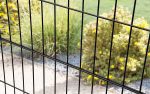 Nærbillede af Metal Hegn "BLACKY" 2D Panel Pulverlakeret Sort Metal, med lodrette og vandrette bjælker, med fokus på stængerne i forgrunden. Bag hegnet er der en have med grønt græs, gule blomstrende buske og en grussti. Baggrunden er lidt ude af fokus.