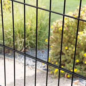 Nærbillede af Metal Hegn "BLACKY" 2D Panel Pulverlakeret Sort Metal, med lodrette og vandrette bjælker, med fokus på stængerne i forgrunden. Bag hegnet er der en have med grønt græs, gule blomstrende buske og en grussti. Baggrunden er lidt ude af fokus.