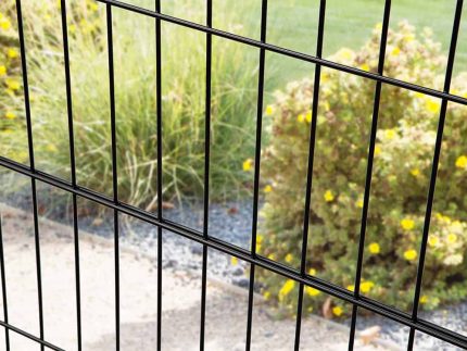 Nærbillede af Metal Hegn "BLACKY" 2D Panel Pulverlakeret Sort Metal, med lodrette og vandrette bjælker, med fokus på stængerne i forgrunden. Bag hegnet er der en have med grønt græs, gule blomstrende buske og en grussti. Baggrunden er lidt ude af fokus.