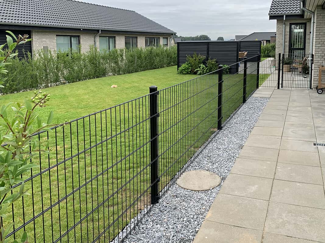 En baggård med en pænt slået græsplæne omgivet af et sort metaltrådshegn. Terrassen har grå belægninger og en grus sektion ved siden af hegnet, suppleret med Havelåge 2D Panelfyld 8/6/8mm Rammeløs "BLACKY" Komplet for nem adgang. Buske fungerer som en privatlivshæk foran nabohuse. Overskyet himmel skaber en rolig, fredfyldt atmosfære.