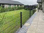En baggård med en pænt slået græsplæne omgivet af et sort metaltrådshegn. Terrassen har grå belægninger og en grus sektion ved siden af hegnet, suppleret med Havelåge 2D Panelfyld 8/6/8mm Rammeløs "BLACKY" Komplet for nem adgang. Buske fungerer som en privatlivshæk foran nabohuse. Overskyet himmel skaber en rolig, fredfyldt atmosfære.