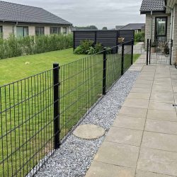 En moderne baghave viser en pænt trimmet græsplæne omkranset af Metal Hegn "BLACKY" 2D Panel Pulverlakeret Sort Metal, komplet med stålstolper og beslag. Terrassen har store firkantede stenfliser, grus langs hegnet, huse med gråt tag i udsigt og buske, der giver grønt.