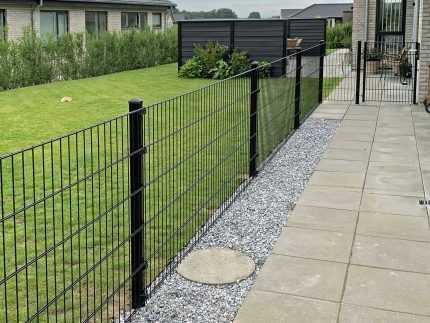 En moderne baghave viser en pænt trimmet græsplæne omkranset af Metal Hegn "BLACKY" 2D Panel Pulverlakeret Sort Metal, komplet med stålstolper og beslag. Terrassen har store firkantede stenfliser, grus langs hegnet, huse med gråt tag i udsigt og buske, der giver grønt.